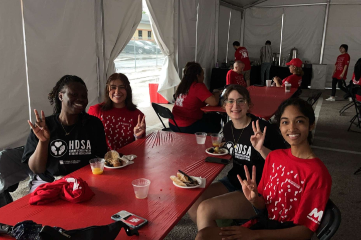 Four University of Houston students volunteer at the 2023 March of Dimes March for Babies event, seated around a red table under a large white tent. They smile and make the 'Go Coogs' hand sign, wearing HDSA and UH volunteer shirts. Plates of breakfast food and drinks are on the table, with other event participants and volunteers visible in the background.