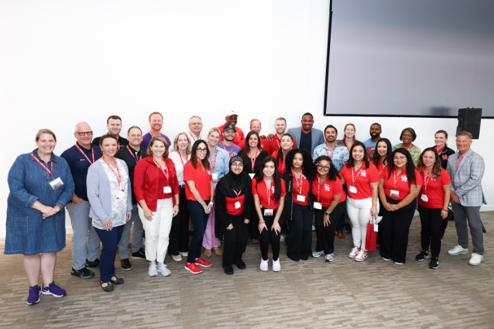 Group photo of University of Houston representatives and attendees at the 2024 Big XII Conference. The group, a mix of students and staff, stands together in a large indoor space, wearing red UH shirts and conference lanyards. They smile and pose in front of a white wall with a large screen partially visible in the background.