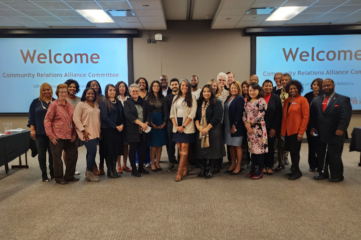 Group photo of the Community Relations Alliance Committee members at the University of Houston. The diverse group stands together in a conference room, smiling in front of a projected screen displaying a welcome message. The committee gathers to foster partnerships and engage with the community, supporting UH’s mission to strengthen community relations.