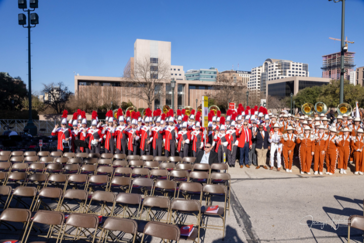 University of Houston band members stand in formation, dressed in red and white uniforms with tall feathered hats, at a government inauguration ceremony. They are positioned in an outdoor venue with rows of empty chairs in the foreground and buildings in the background, prepared to perform and represent UH at the event.