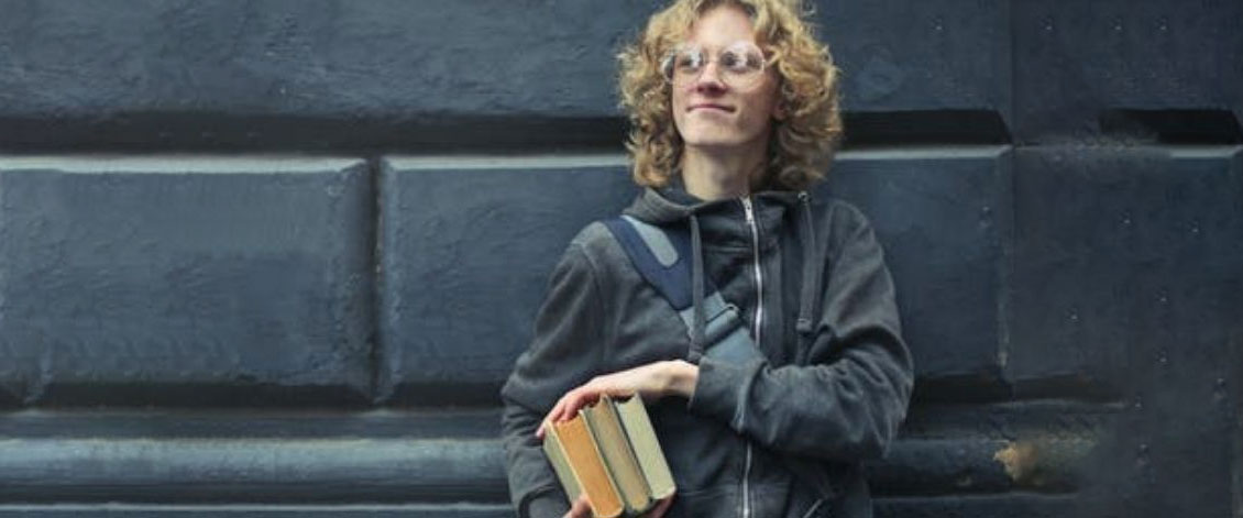 A young man holding 3 books and looking up while reclined on a brick wall