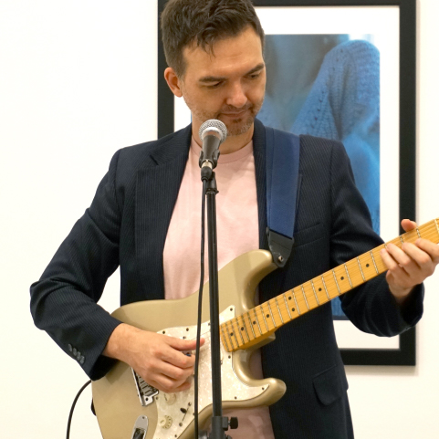 Close up of a performer with cropped dark hair, wearing a dark blazer and pale t-shirt behind a microphone playing an electric guitar.