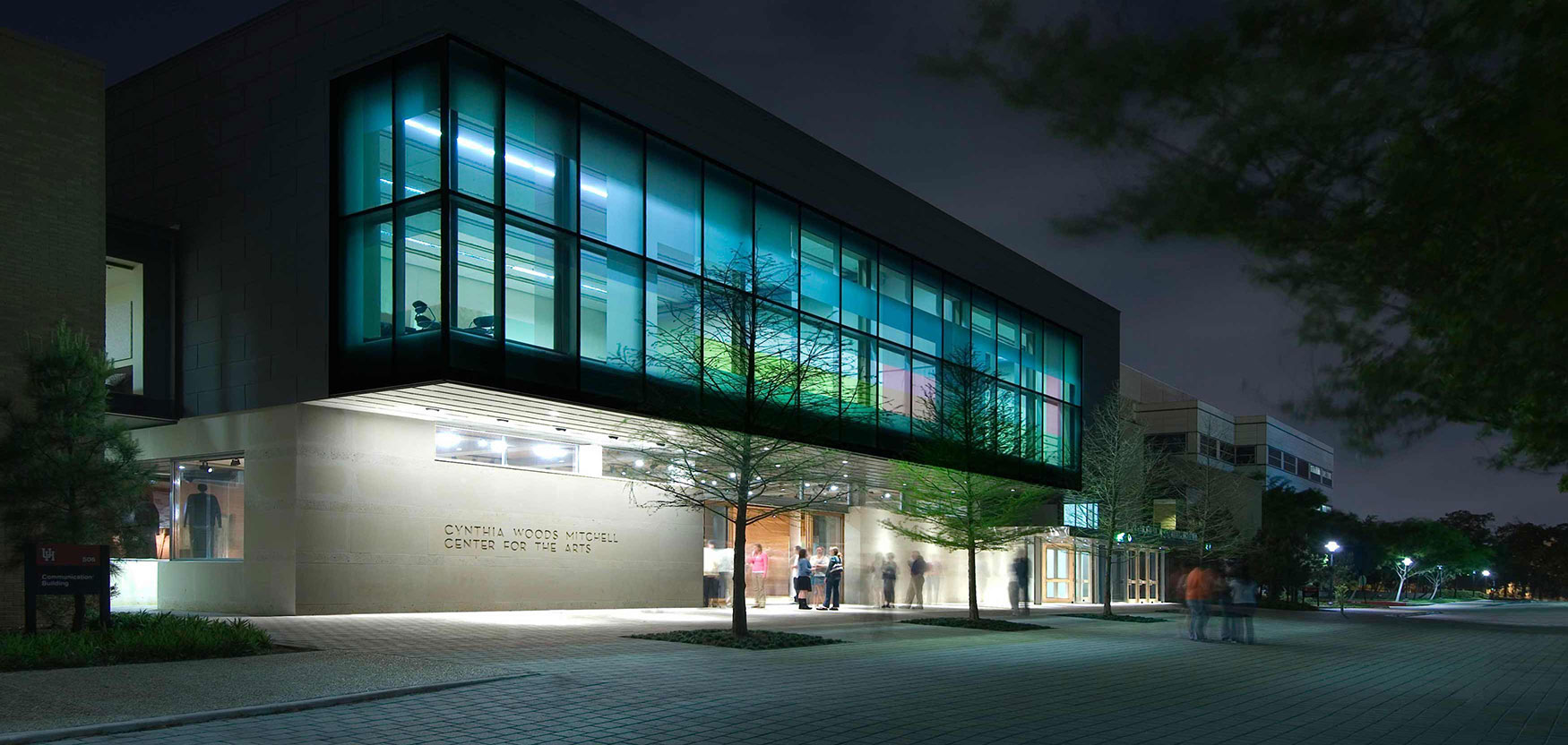 A building lit up at night with ‘Cynthia Woods Mitchell Center for the Arts’ inscribed on the cream stone façade of the ground floor and dark glass on the second floor and people are gathered near the lit entrance.