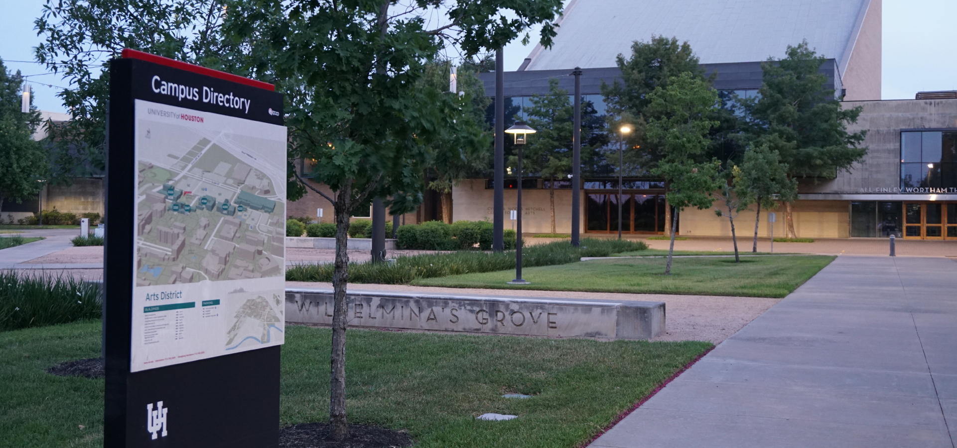 The University of Houston’s arts district campus signage stands in front of a grassy courtyard and a concrete bench engraved with “Wilhelmina’s Grove,” the cream and grey Mitchell Center building visible between trees in the background.