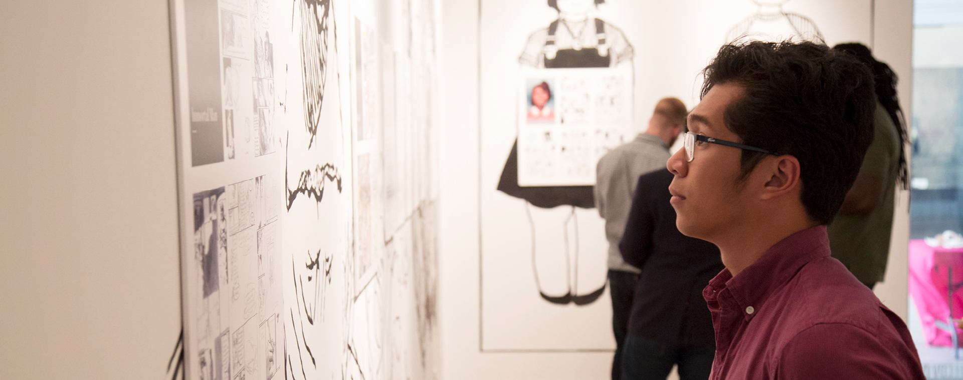 A person with short dark hair, wearing glasses and a maroon collared shirt stands in profile looking at artwork on the wall, other people stand around in the background.