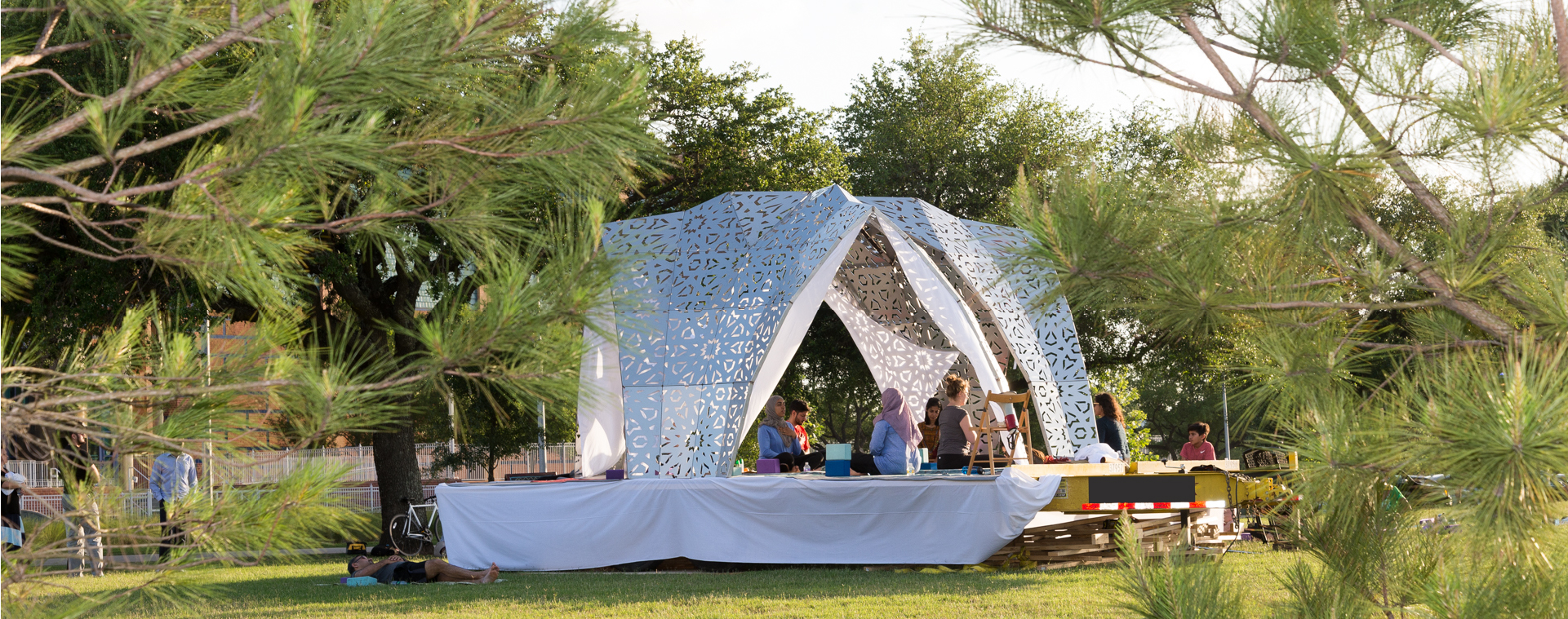 People meditating in the shade of metal architectural dome-like structure with four arched entrances and geometric cutout patterns on a platform in a lush park.