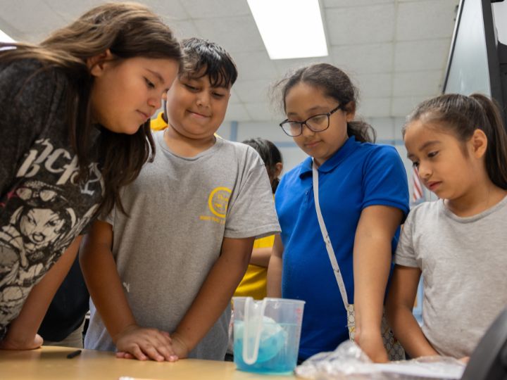 Four students around a table, focused on a science experiment involving a beaker and blue liquid.