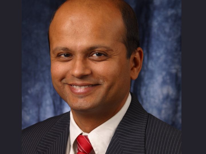 Portrait of Bhavin Sheth wearing a formal pinstripe suit with a red tie, smiling against a mottled blue backdrop.