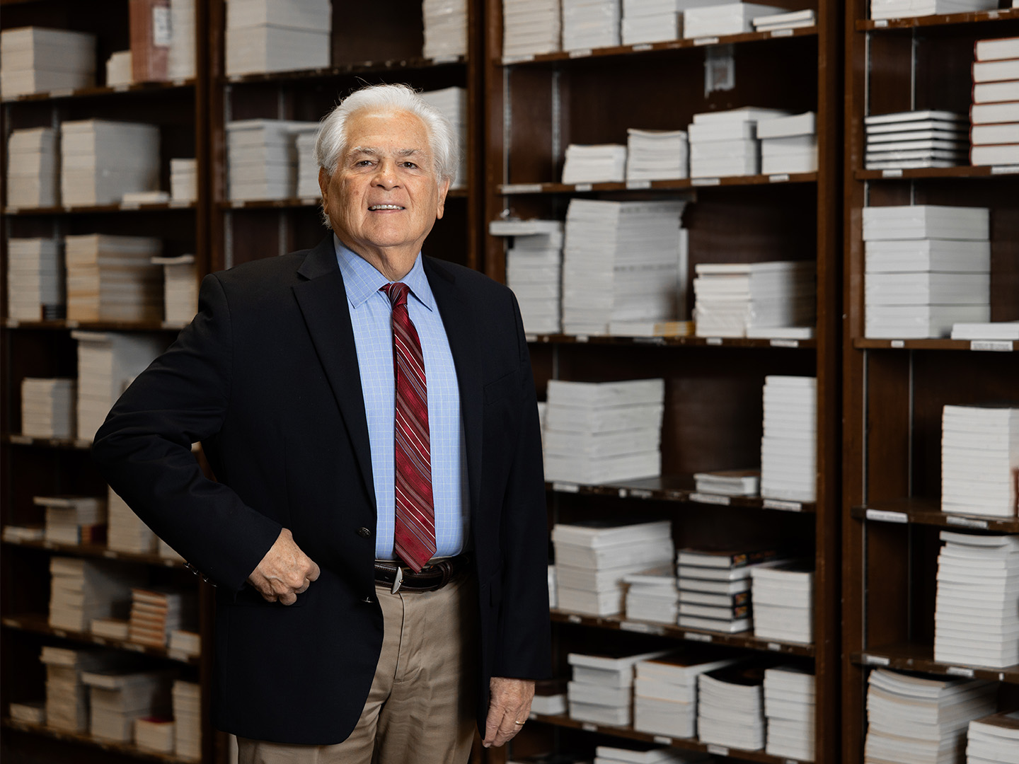 UH’s Nicolás Kanellos standing in a room with shelves stocked with many books, smiling at the camera. He’s dressed in a business suit and red tie.