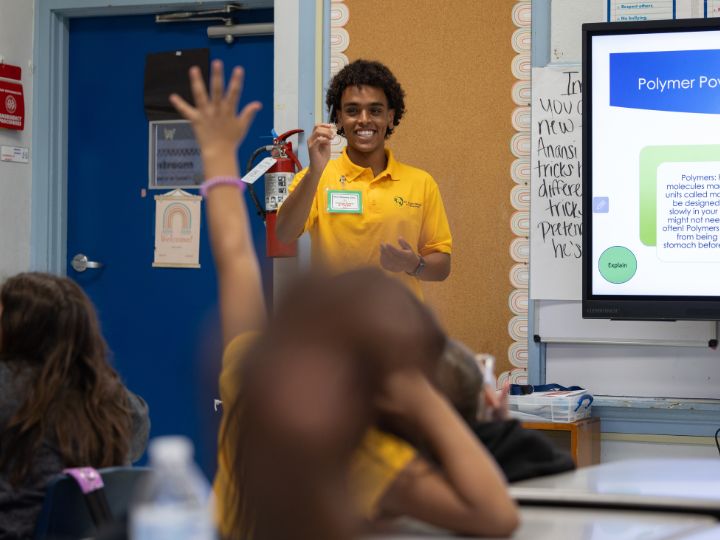 Teacher smiling while holding up a model to a classroom of engaged students, with a presentation visible on the screen behind.