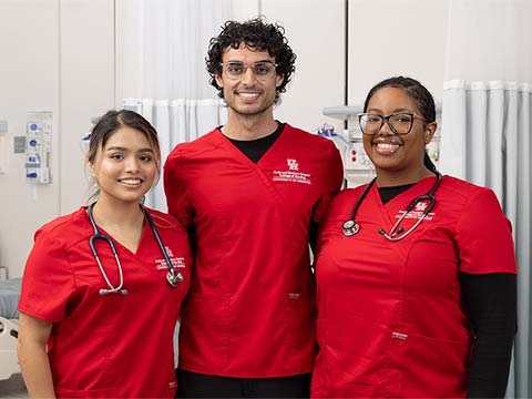 Three nursing students in red scrubs stand together in front of hospital beds.